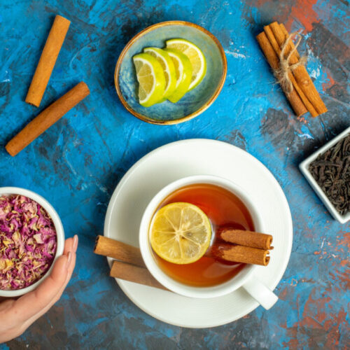 top view cup tea with lemon cinnamon sticks woman hand holding dried rose petals bowl blue red surface 1024x683 1